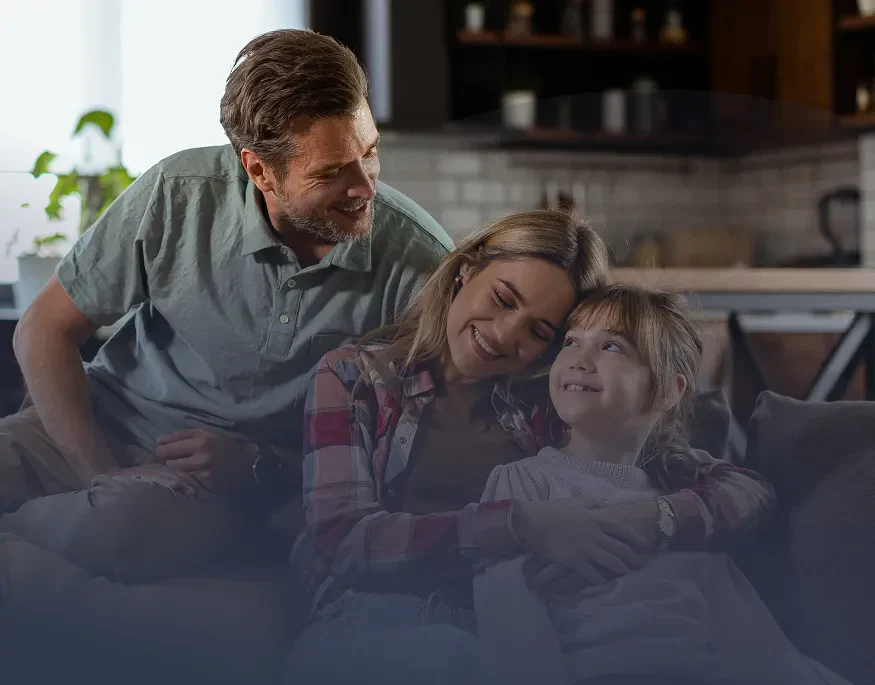 A family of three sits together on a couch, enjoying a cozy moment in a warmly lit living space with a plant in the background.