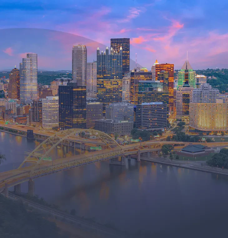 Panoramic view of Pittsburgh's skyline at dusk, featuring illuminated skyscrapers, a bridge, and the winding river below.