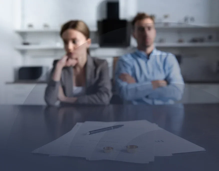 A couple sits with crossed arms, looking serious, beside divorce papers and wedding rings on a table in a softly lit room.