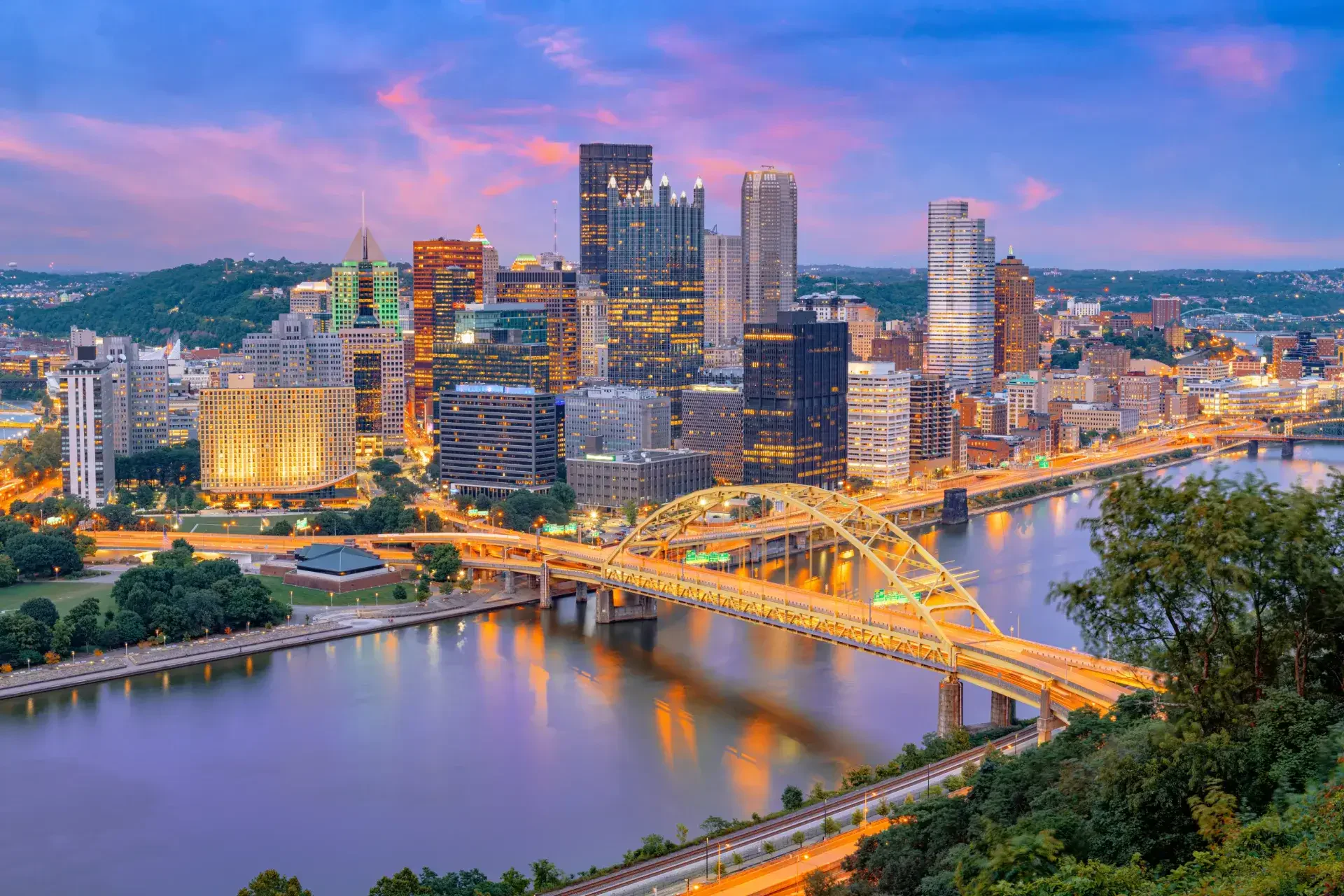 Pittsburgh city skyline at dusk with illuminated buildings, bridges, and rivers visible under a colorful sky.