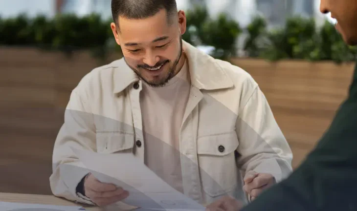 A person in a light jacket reviews documents at a table while another person gestures, discussing the materials in a casual setting.