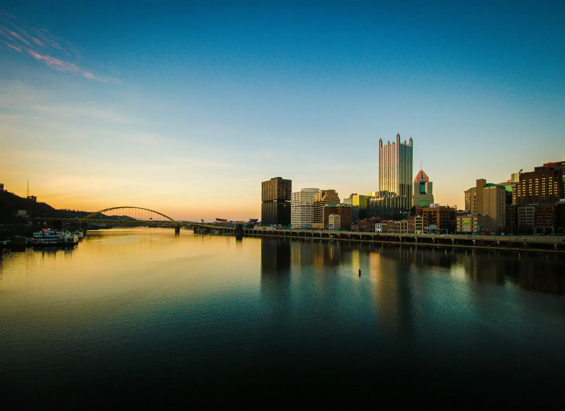 Sunset over Pittsburgh skyline reflecting in the river, featuring iconic buildings and a bridge against a clear blue sky.