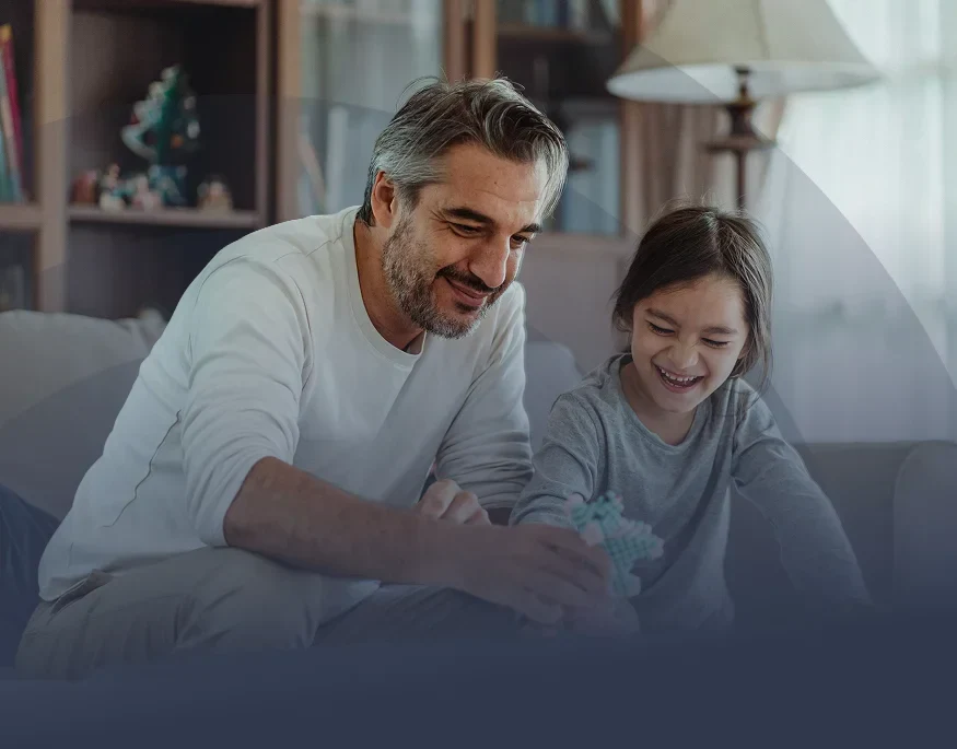 A child and an adult sit together on a couch, engaged in play with a colorful toy in a cozy, well-lit living room.